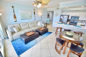 A living room with a sofa, ottomans, and a dining table with chairs. There is a kitchen area with appliances and cabinets. - Destin West Gulfside #411 ~Free Beach Service! ~Great View! Pools + Lazy River! (Fort Walton Beach)