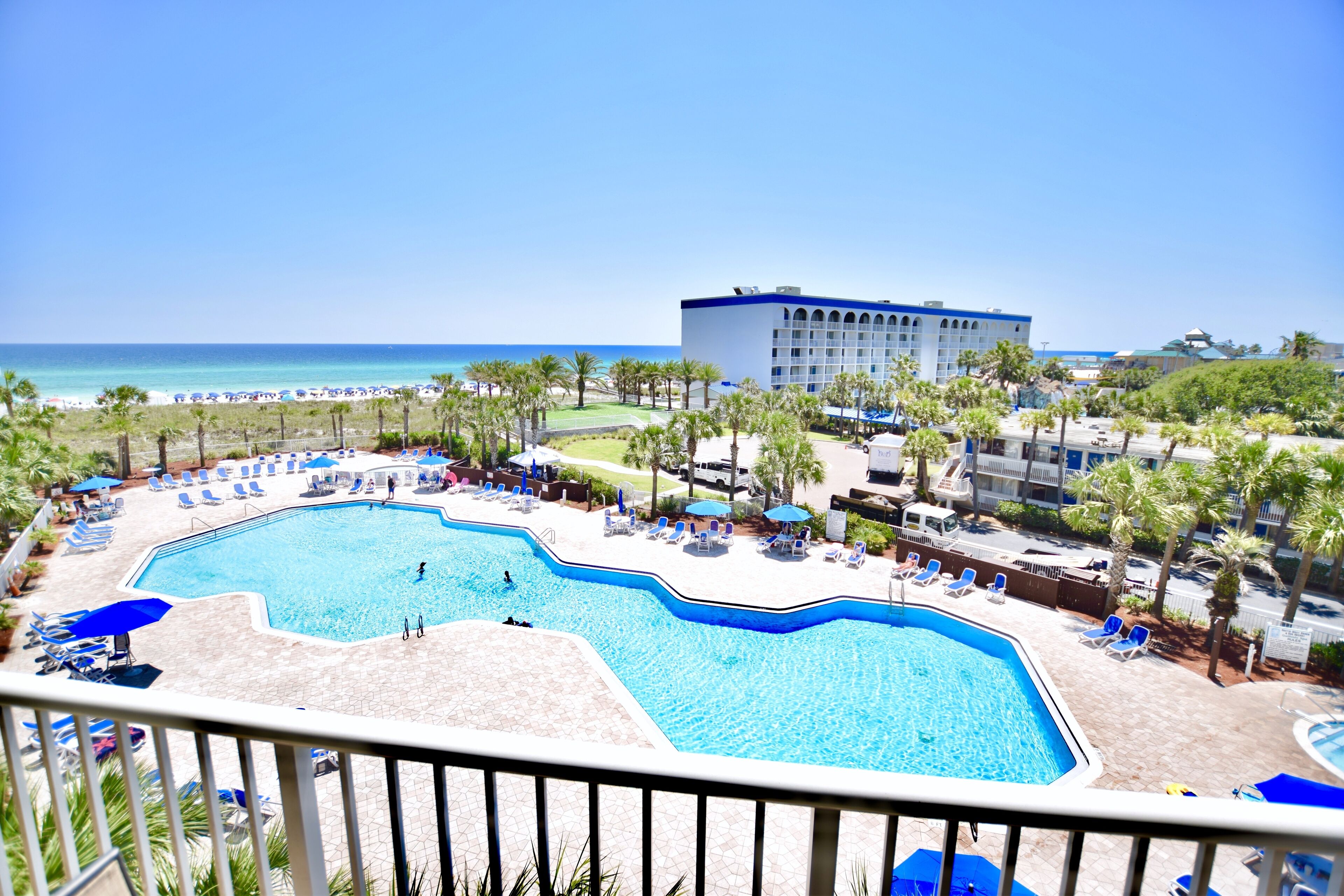 A pool area with lounge chairs and umbrellas, a building in the background, and a view of the beach.