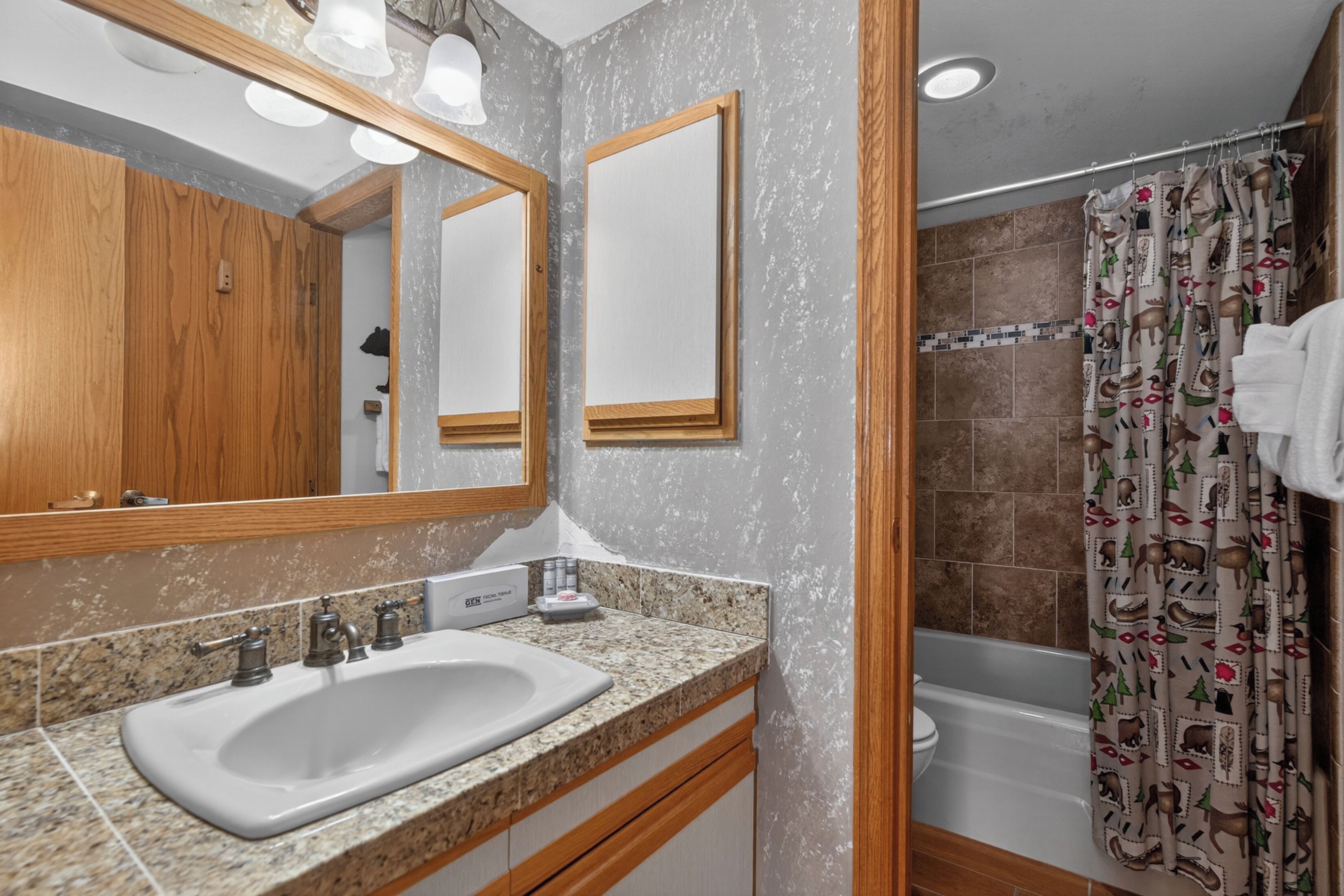 A bathroom with a granite countertop sink, wooden cabinets, a large mirror, and a shower curtain with a deer pattern.