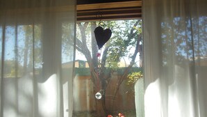 A window with a heart-shaped ornament hanging in the center, surrounded by a garden with red flowers and trees.