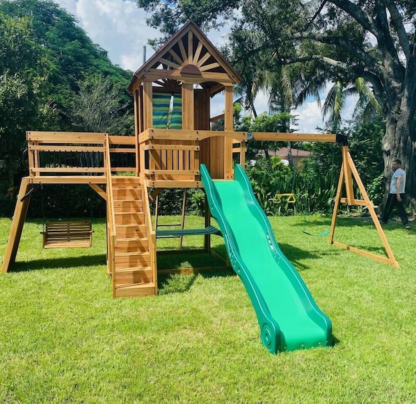 Una struttura giochi in legno con scivolo, scaletta e una piccola casetta.