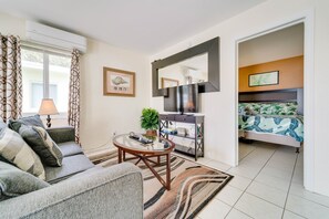 A modern living room with a sofa, coffee table, and TV stand. There is a bed with a patterned bedspread in the adjacent room. - #3 Heated Pool, Large Yard, 1BR+sunroom/office (Fort Lauderdale)