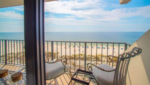 A balcony with two chairs and a small table, overlooking a beach with people and umbrellas. - Beautiful direct Gulf view & beach front condo. (Gulf Shores)
