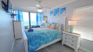 Une chambre à coucher comprenant un lit, une table de chevet, un ventilateur de plafond, une fenêtre avec des rideaux et des œuvres d’art accrochées au mur.