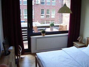 A hotel room with a bed, a window with curtains, a radiator, a small table with a lamp, and a view of a brick building outside.