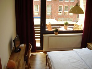 A bedroom with a bed, a desk, a chair, a window with a view of a brick building, and a potted plant on the windowsill.