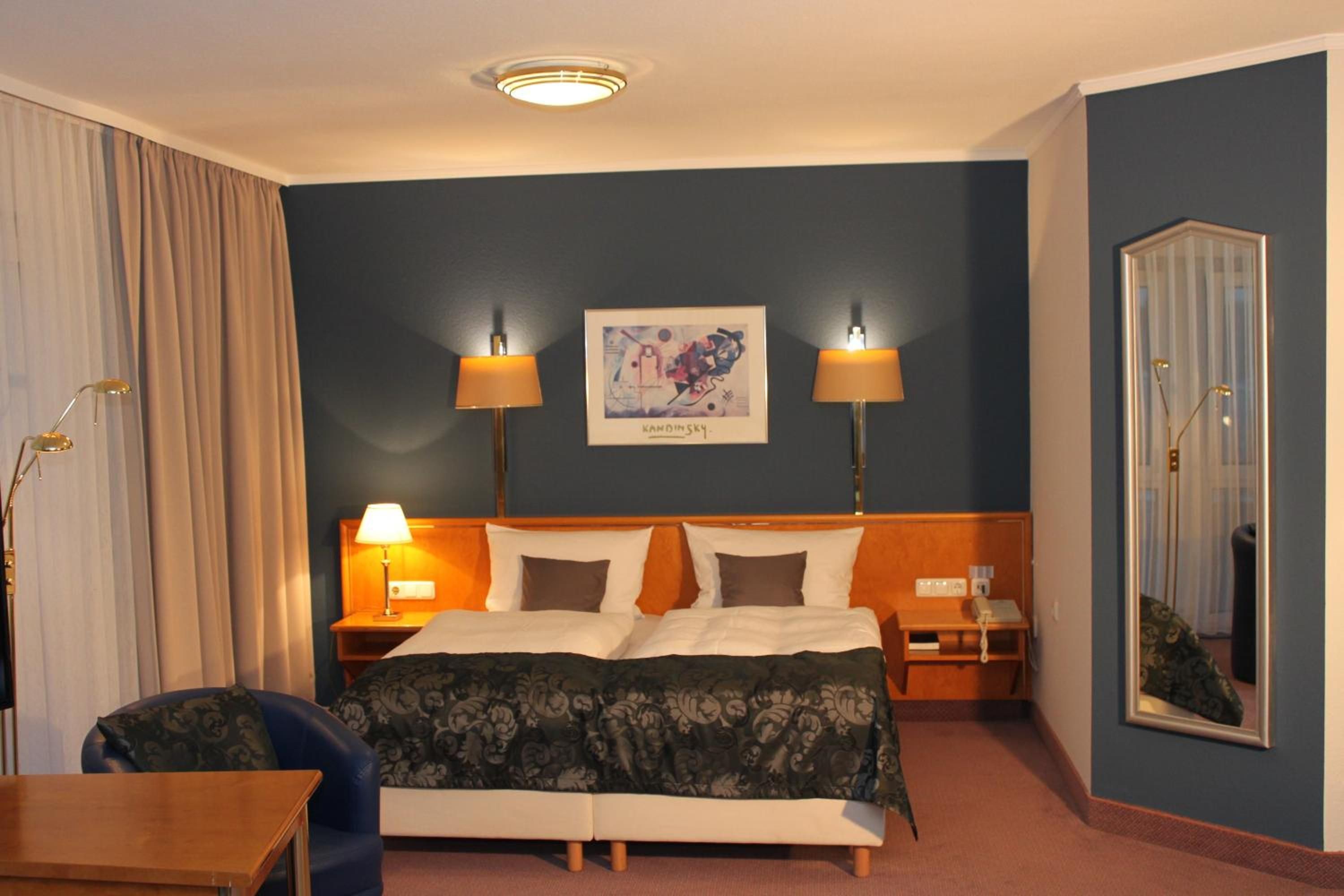 Ein Hotelzimmer mit einem Bett, zwei Nachttischlampen, einem gerahmten Kunstwerk, einem Nachttisch, einem Sessel und einem Spiegel.