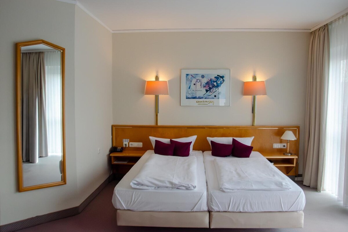 A hotel room with two beds, a large mirror, two bedside tables with lamps, a framed artwork on the wall, and a window with curtains.