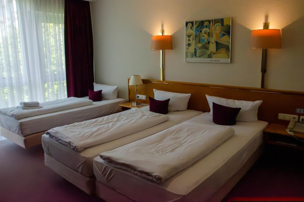 A hotel room with two single beds, a nightstand with a lamp, a painting on the wall, and a window with curtains.