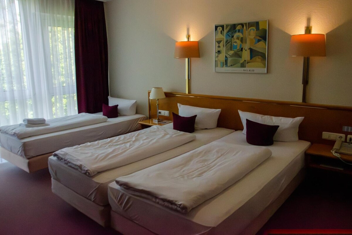 Quarto de hotel com duas camas de solteiro, mesa de cabeceira com luminária, quadro na parede e janela com cortinas.