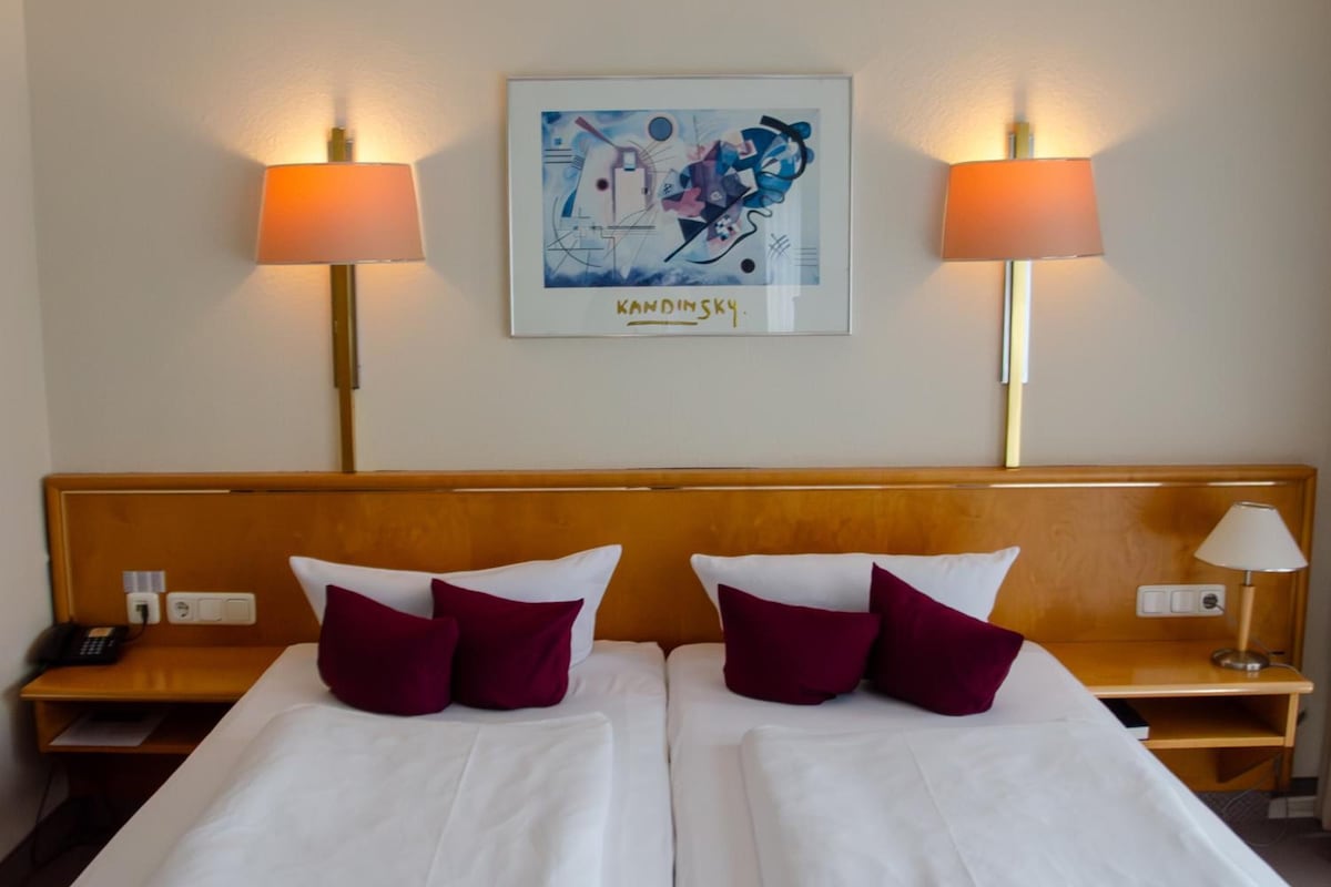 Quarto de hotel com duas camas, cada uma com lençóis brancos e almofadas bordô, cabeceira de madeira, duas luminárias de cabeceira, uma obra de arte emoldurada na parede e uma mesa de cabeceira com telefone e luminária.
