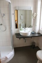 Un baño con ducha envuelta en vidrio, un lavamanos blanco sobre encimera negra, un espejo sobre el lavamanos y un inodoro.