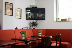 A room with a red sofa, two tables with chairs, a television mounted on the wall, and a window with a view of the city. - Journal Cityhotel (Wolfsburg)