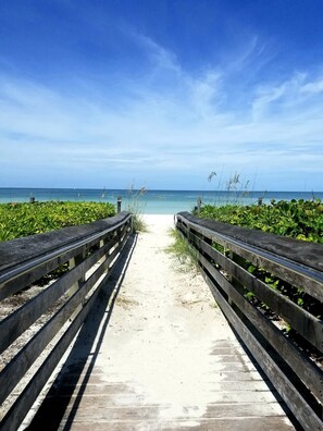 A wooden boardwalk leading to a beach with clear blue water and a sandy path.