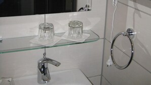 Baño con espejo, dos secadores de pelo, repisa de cristal con dos vasos, lavabo y un aro para toallas.