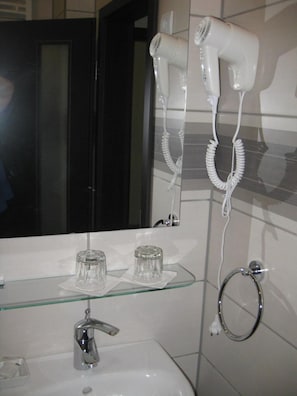 A bathroom with a mirror, two hair dryers, a glass shelf with two glasses, a sink, and a towel ring.
