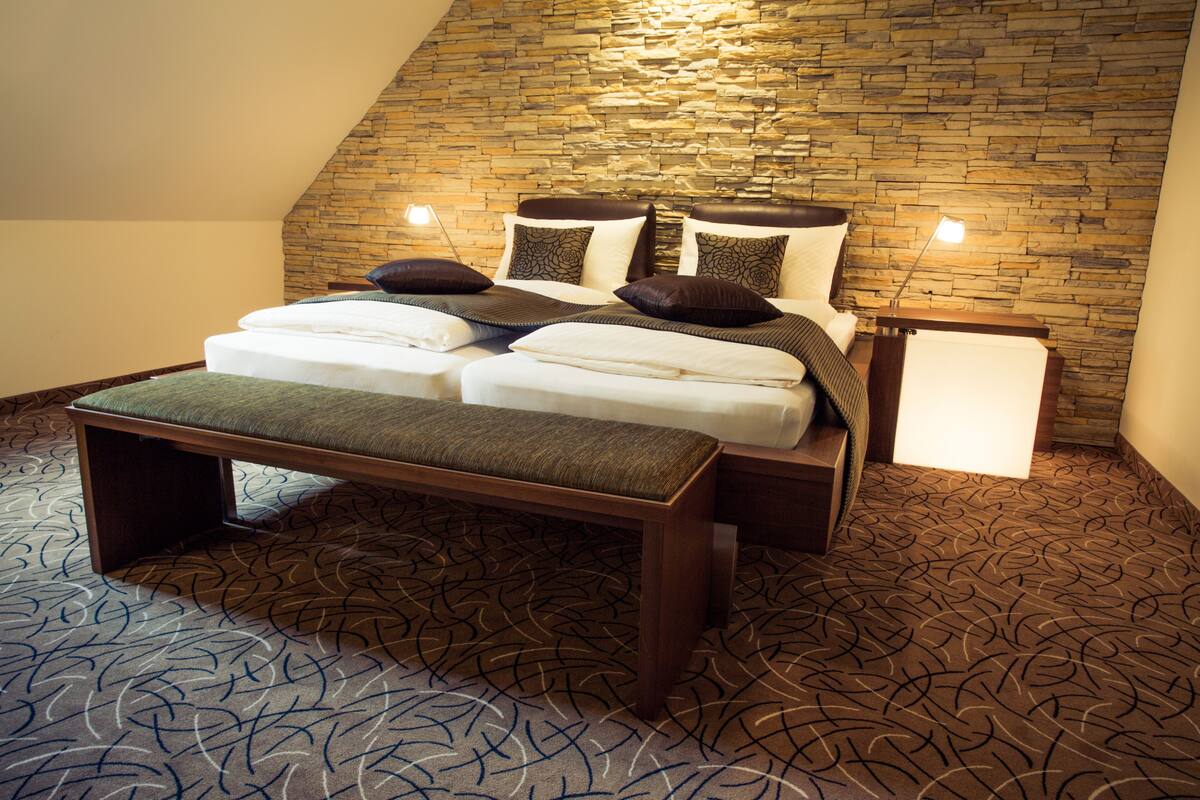 Une chambre avec un mur en pierre, un lit avec de nombreux oreillers, un banc et une table de chevet avec une lampe.
