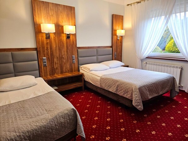Quarto de hotel com duas camas, cabeceira de madeira, tapete vermelho e janela com cortinas.