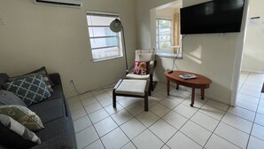 A living room with a sofa, a chair, a small table, and a TV mounted on the wall. - #6 Heated Pool, Private Patio & Grill, 1BR+sunroom (Fort Lauderdale)