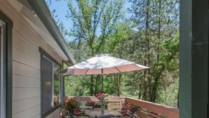 A wooden deck with a table and chairs under a patio umbrella, surrounded by potted plants and a wooden railing.