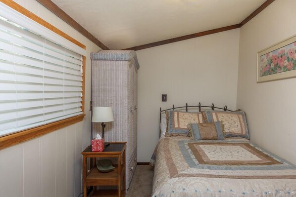 A bedroom with a bed, a bedside table with a lamp, a window with blinds, and a painting on the wall.