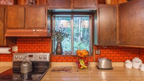A kitchen with a brick backsplash, wooden cabinets, a stove, a window with a view of trees, and various kitchen items on the counter.