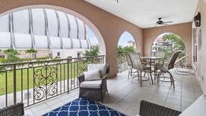 A covered balcony with a view of a modern building, a dining area with chairs and a table, and a ceiling fan.