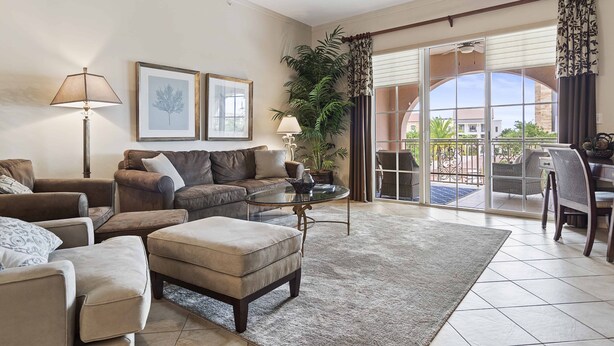 A spacious living room with a sofa, ottoman, and a glass coffee table. There is a ceiling fan, a potted plant, and a view of the outside through large sliding glass doors.