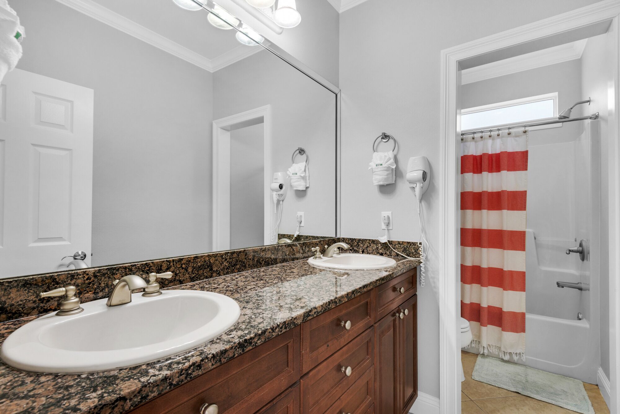 A bathroom with two sinks, a vanity with drawers, a mirror, a hairdryer, and a shower with a striped curtain.