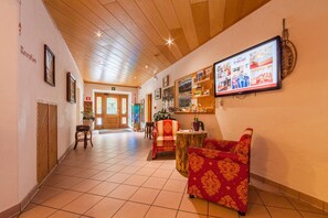 A hotel lobby with a wooden ceiling, a reception desk, a seating area with red upholstered chairs, and a wall-mounted TV displaying hotel information. - Hotel Tolderhof (San Candido)