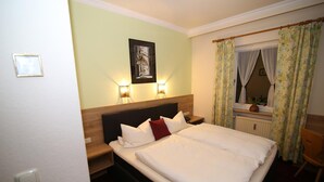 Habitación de hotel con cama, dos lámparas de noche, una mesita auxiliar, cabecero de madera, un cuadro enmarcado en la pared y cortinas con estampado floral.