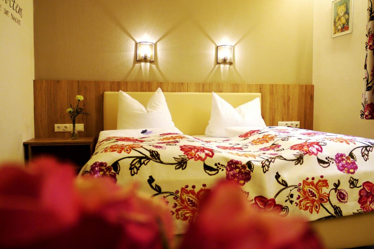 A hotel room with a bed, two wall-mounted lights, a nightstand, and a floral bedspread.