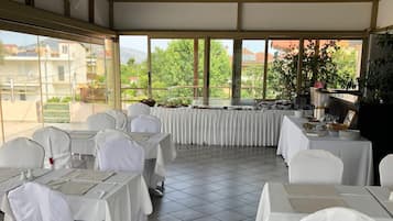 Un espace de restauration extérieur abrité, doté de tables et de chaises, de grandes fenêtres et offrant une vue sur les bâtiments et la verdure.