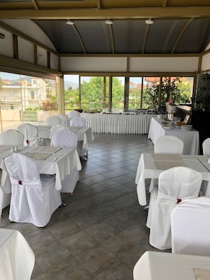 A covered outdoor dining area with tables and chairs, large windows, and a view of buildings and greenery. - Selefkos Palace (Igoumenitsa)