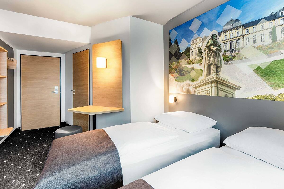 Hotellihuone, jossa on kaksi sänkyä, puuovi, pieni pöytä ja seinään kiinnitetty valaisin.
