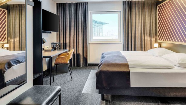 A hotel room with a bed, a desk, a chair, a large mirror, and a window with a view of a building. - B&B Hotel Bonn-West (Bonn)