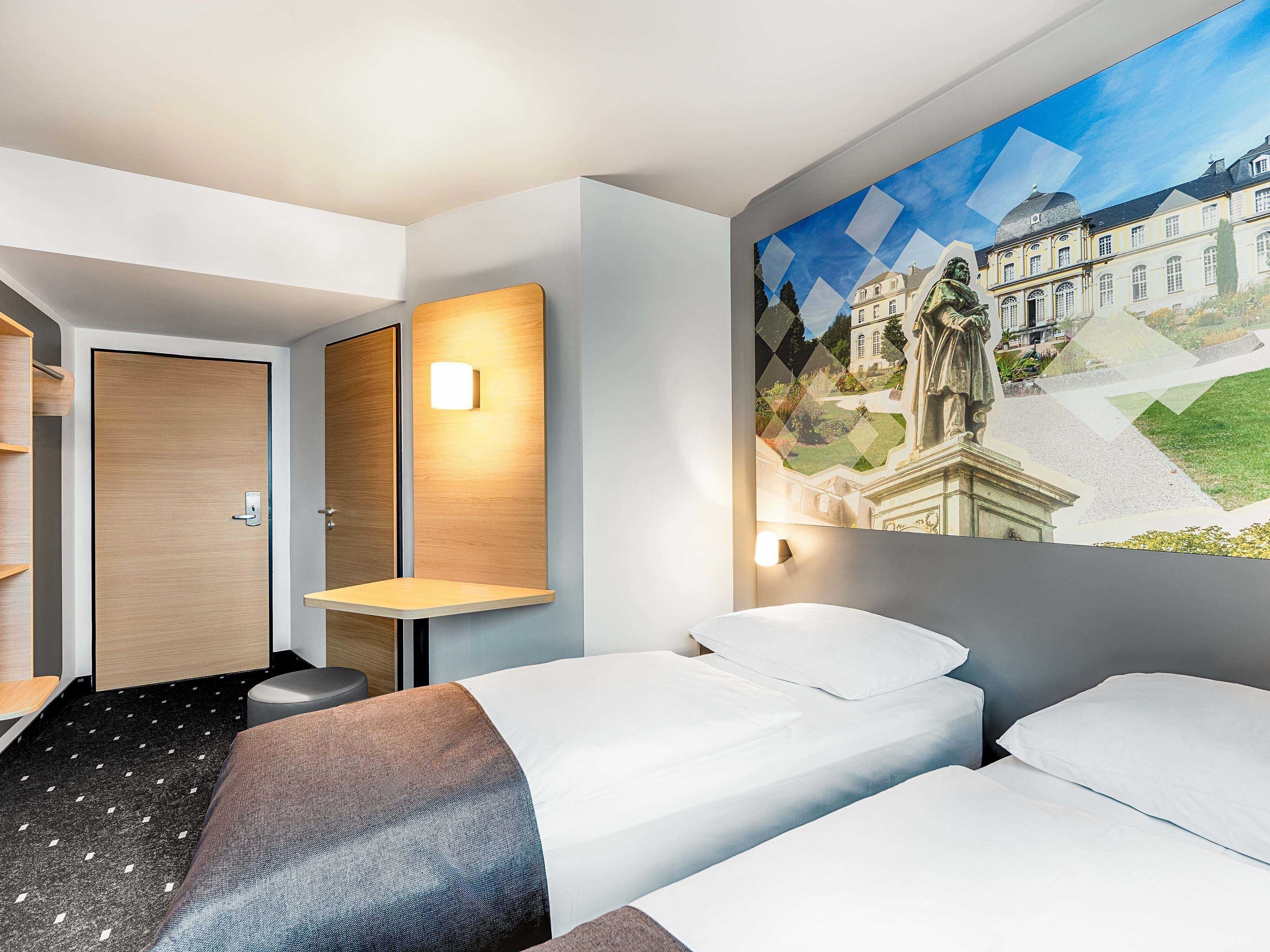 Habitación de hotel con dos camas, una puerta de madera, una mesita y una lámpara fijada a la pared.