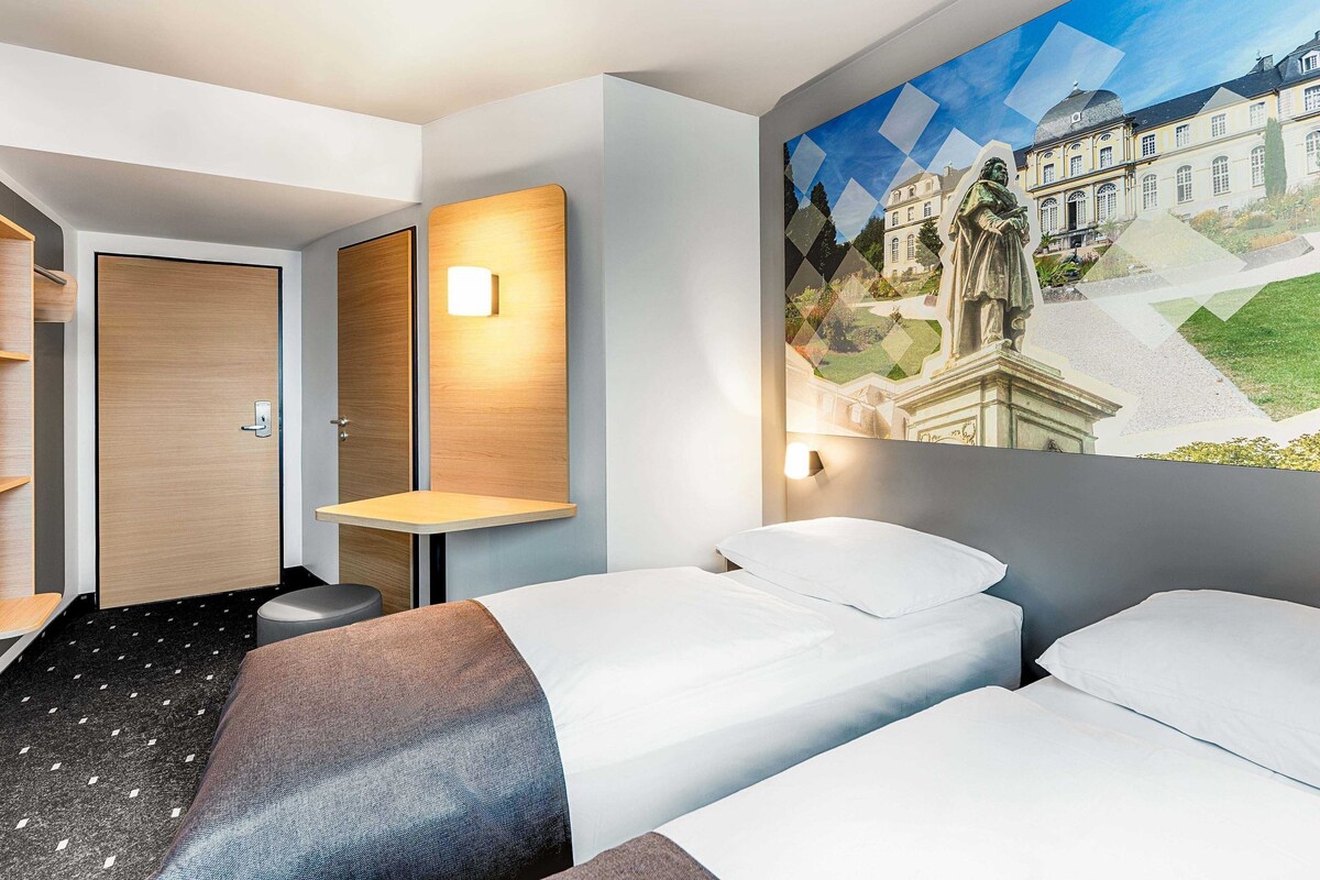 A hotel room with two beds, a wooden door, a small table, and a wall-mounted light.