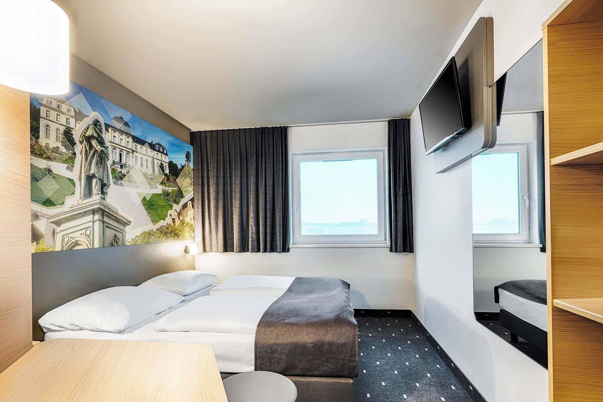 Hotellihuone, jossa on kaksi sänkyä, työpöytä, televisio ja maalaus, jossa on kuvattuna rakennus.