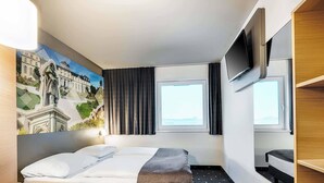 A hotel room with two beds, a desk, a TV, and a painting of a building. - B&B Hotel Bonn-West (Bonn)