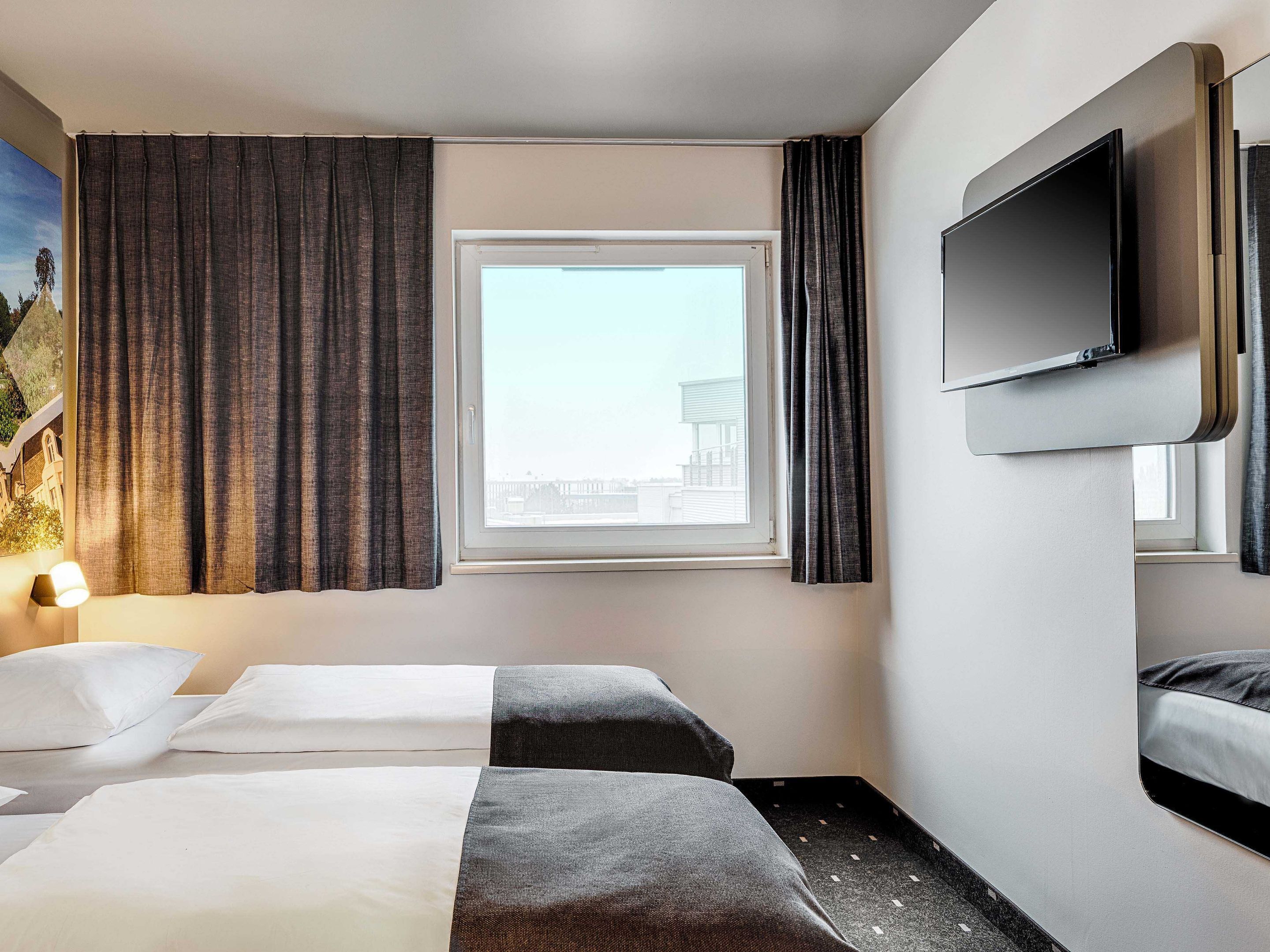 A hotel room with two beds, a TV mounted on the wall, a window with curtains, and a painting on the wall.