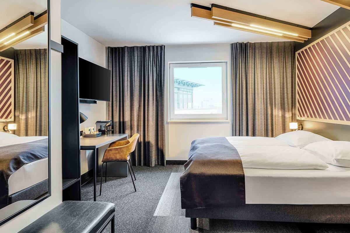 Hotellihuone, jossa on sänky, työpöytä, tuoli, suuri peili ja ikkuna, josta on näkymä rakennukseen.
