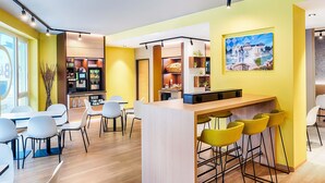 A modern cafe with a yellow and white color scheme, featuring a bar area with yellow stools, white chairs, and a wooden counter. - B&B Hotel Bonn-West (Bonn)