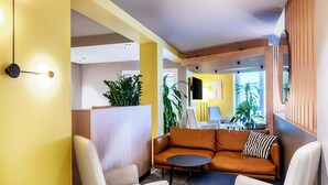 A modern lounge area with a yellow accent wall, a brown leather sofa, white armchairs, and a small round table. - B&B HOTEL Bonn-West (Bonn)
