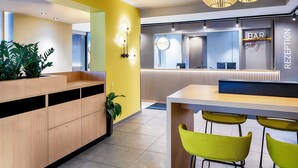A modern reception area with a yellow accent wall, a wooden reception desk, and a bar sign. - B&B Hotel Bonn-West (Bonn)