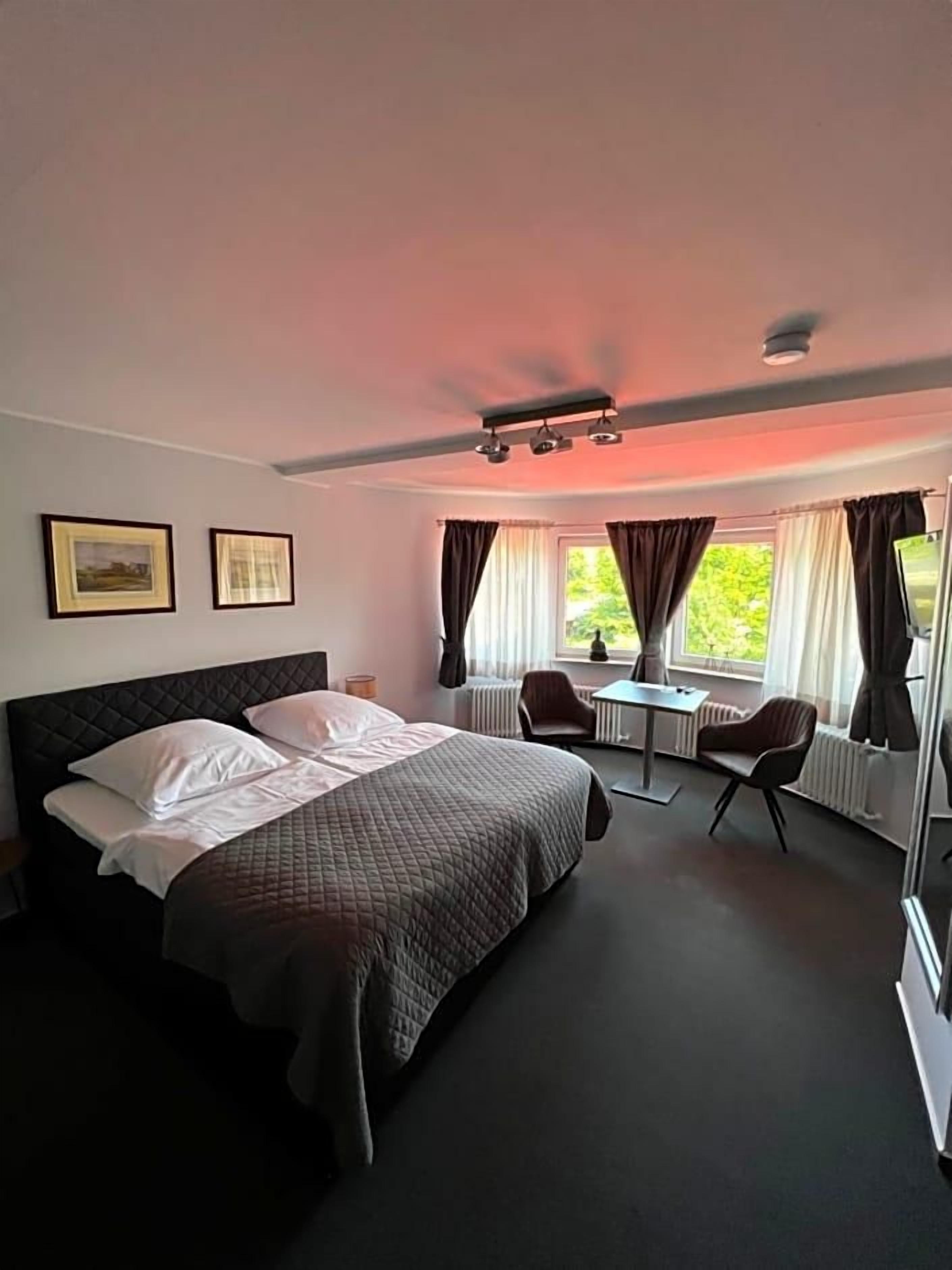 Habitación de hotel con cama, escritorio, dos sillas, ventana con cortinas y una lámpara en el techo.