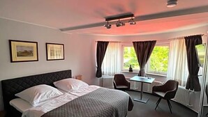 A hotel room with a bed, a desk, two chairs, a window with curtains, and a ceiling light fixture.
