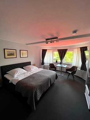 A hotel room with a bed, a desk, two chairs, a window with curtains, and a ceiling light fixture.