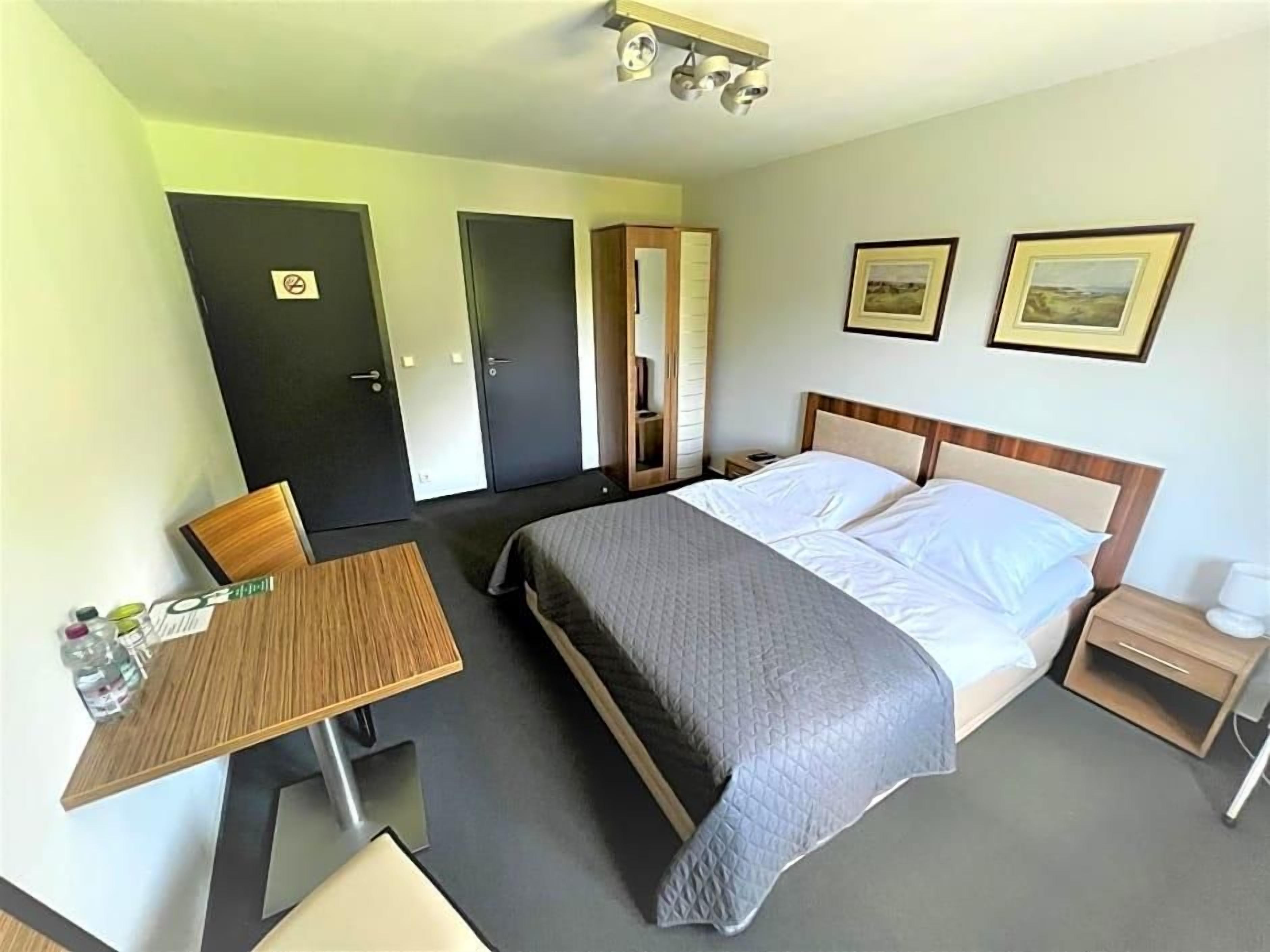 A hotel room with a bed, bedside table, chair, and a desk with a chair. There are two doors, one with a 'No Entry' sign, and two framed pictures on the wall.