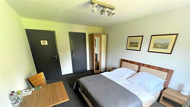 A hotel room with a bed, bedside table, chair, and a desk with a chair. There are two doors, one with a 'No Entry' sign, and two framed pictures on the wall.